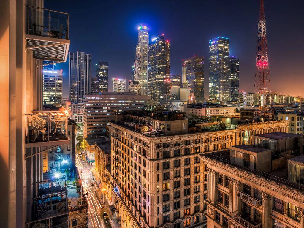 Na obrázku je zachytená panoráma Los Angeles, jedného z najznámejších a najživších miest v USA. V popredí vidíme mestskú krajinu s modernými mrakodrapmi, ktoré sa týčia nad mestom, symbolizujúce jeho obchodný a finančný význam. V pozadí je zreteľná silueta hôr, ktoré obklopujú mesto, a jasné modré nebo typické pre kalifornské podnebie. Slnečné svetlo osvetľuje scenériu, čo dodáva mestu charakteristickú teplú a živú atmosféru. Mesto je známe svojím kultúrnym bohatstvom, rozmanitosťou a ikonickými pamiatkami, ako sú Hollywood, pláže Santa Monica a Venice, alebo štúdiá Universal Studios. Na uliciach mesta je stále rušno, s autami a chodcami, ktoré prispievajú k neustálemu pohybu Los Angeles. Mesto je synonymom pre filmový priemysel a zábavu, pričom ponúka nekonečné možnosti pre turistov aj obyvateľov, ktorí hľadajú zábavu, kultúru a príležitosti.