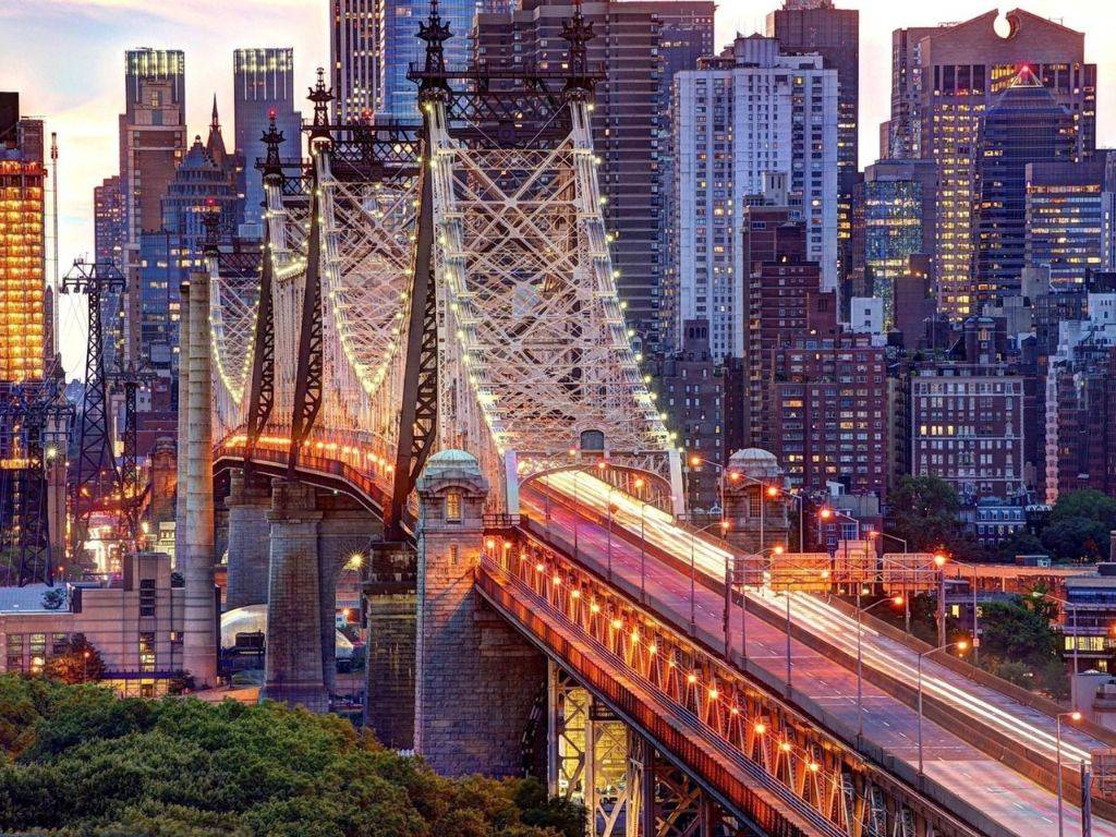 Na obrázku je zachytený Queensboro Bridge, elegantný a technicky impozantný most, ktorý spája Manhattan a Queens cez rieku East River v New Yorku. Most, známy aj ako 59th Street Bridge, je známy svojím charakteristickým oceľovým rámom a výrazným dizajnom, ktorý vytvára robustný, ale zároveň esteticky príťažlivý vzhľad. Základné štruktúry mosta zahŕňajú zvislé piliere a horizontálne nosníky, ktoré mu dodávajú stabilitu a zároveň krásne prepojenie medzi oboma časťami mesta. V pozadí je panoráma Manhattanu, plná vysokých budov, ktoré tvoria moderný mestský horizont. East River pod mostom je pokojná, s občasným pohybom lodí a člnov. Queensboro Bridge je dôležitým dopravným spojom a zároveň jedným z najikonickejších mostov v New Yorku, ponúkajúc nielen praktické využitie, ale aj krásne výhľady na mesto, ktoré obklopuje.